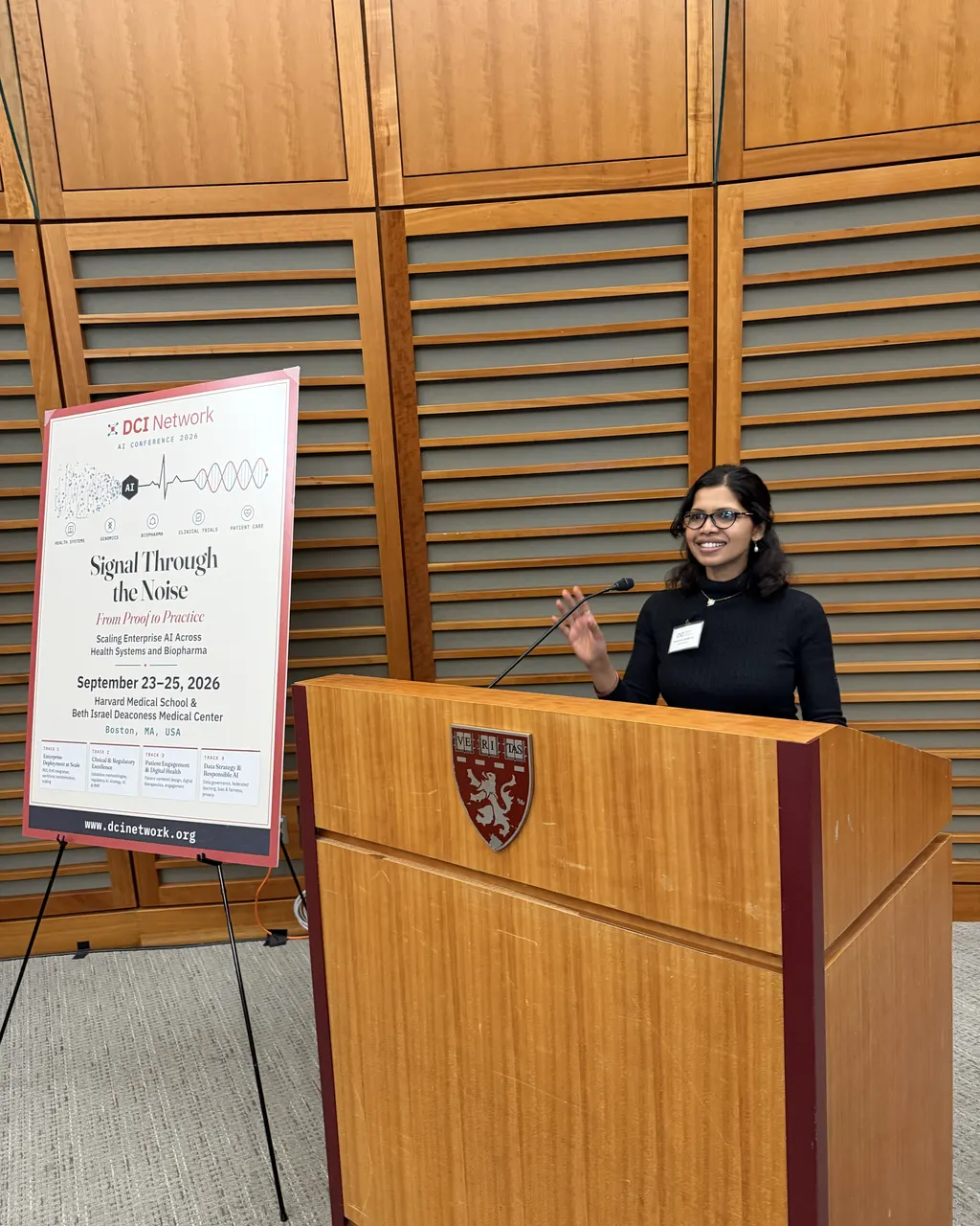 A speaker at a podium at the DCI Network AI Conference 2026, beside the conference poster reading Signal Through the Noise